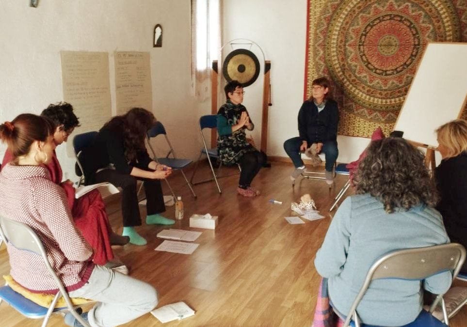 Un pequeño grupo de personas sentadas en círculo en sillas y cojines en una sala, enfrascadas en una discusión. En el suelo hay papeles y una botella de agua; al fondo, un tapiz y un rotafolio.