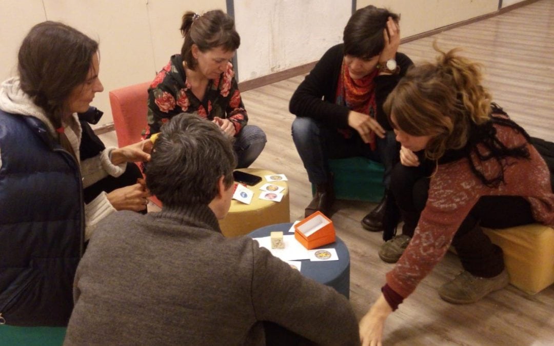 Cinco personas se sientan en círculo en asientos bajos y participan en un juego o actividad de mesa con cartas y pequeños objetos sobre una mesa.