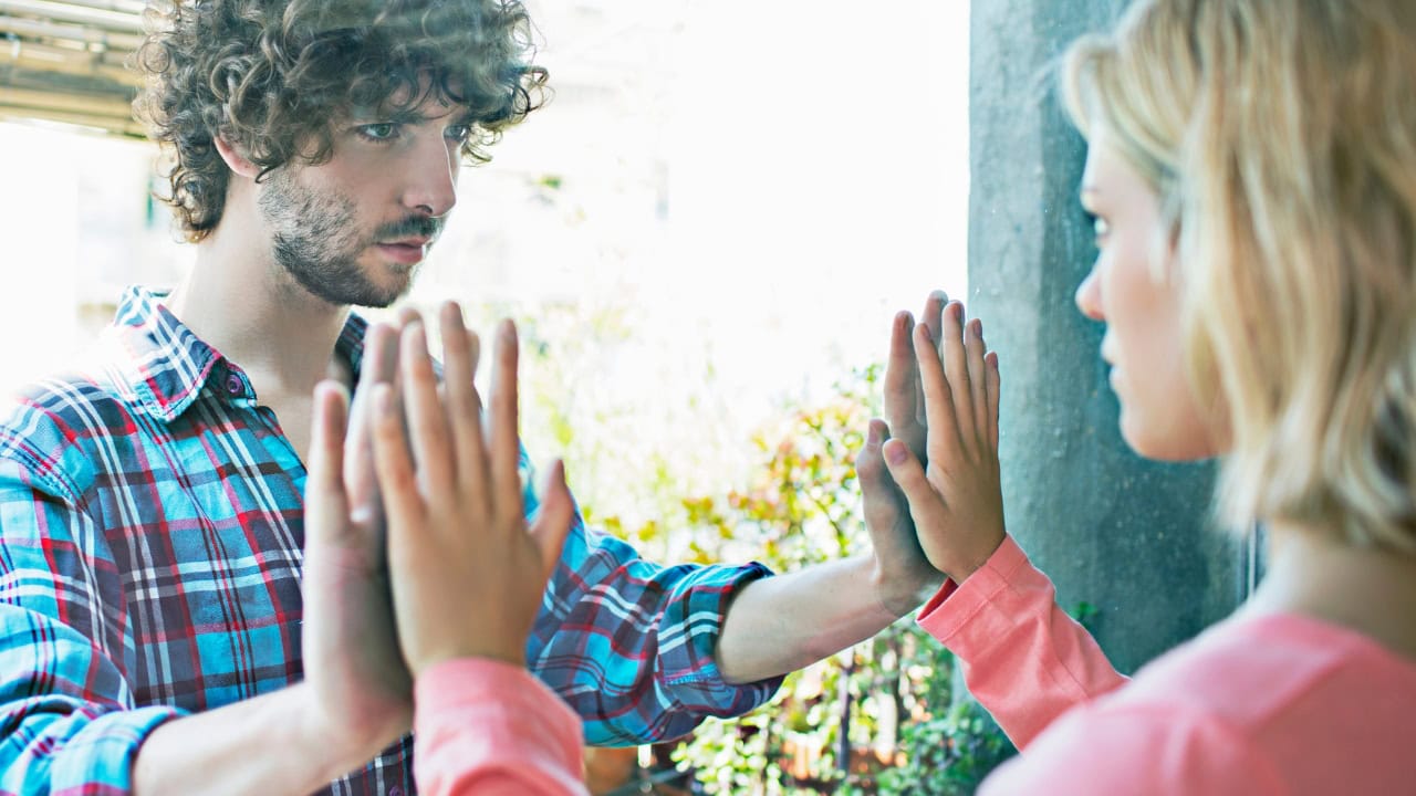 Un chico y una chica se miran con gesto serio mientras juntan las palmas de sus manos en un cristal que los separa