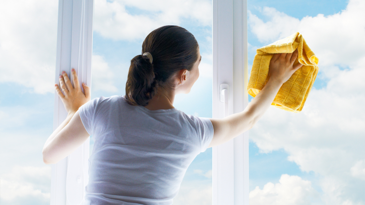 mujer de espaldas limpiando un cristal, la parte limpia deja ver un cielo azul con nubes blancas