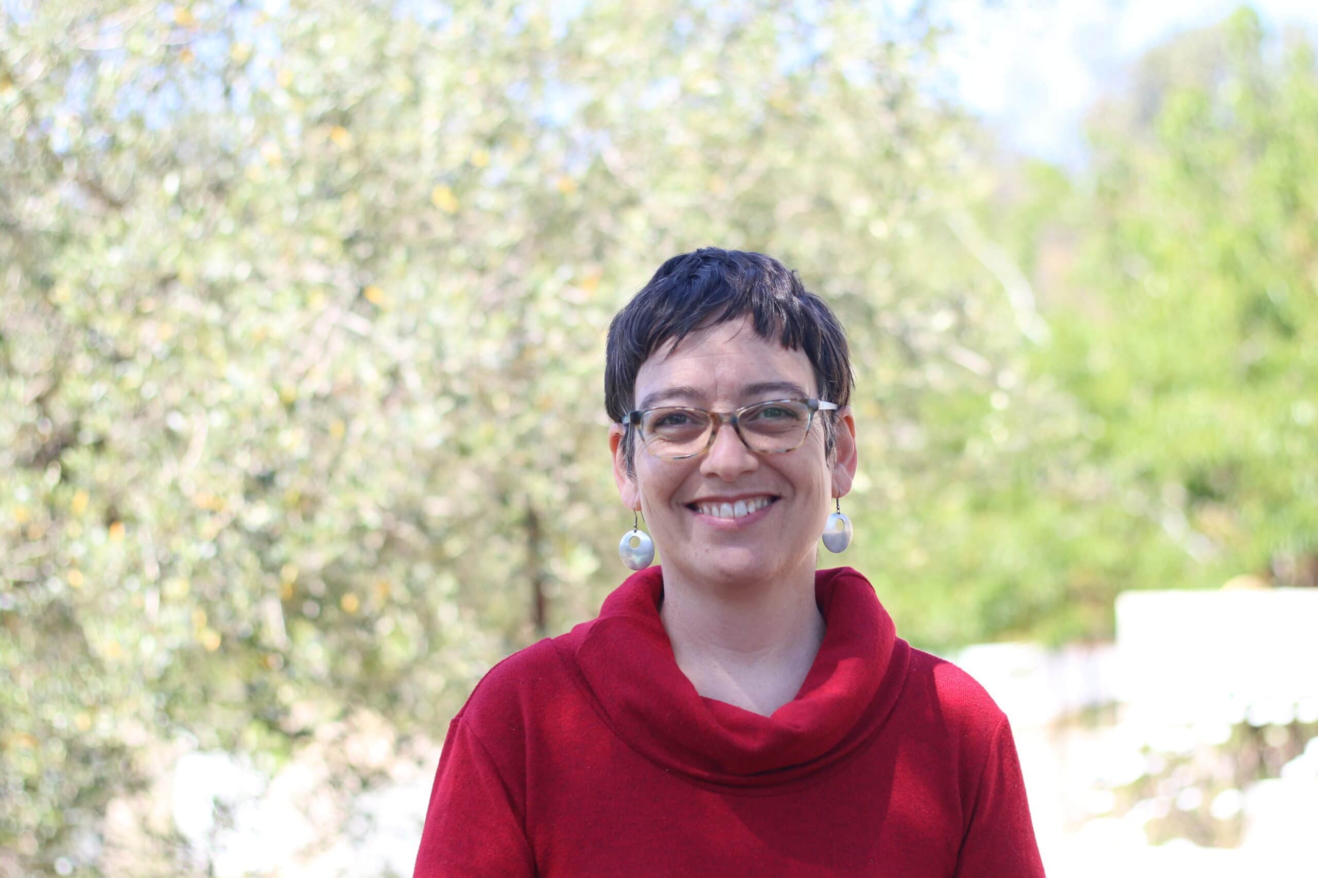 Una persona con pelo corto y oscuro, gafas y pendientes sonríe al aire libre, llevando un jersey rojo. Al fondo se ven árboles y vegetación. Una persona con pelo corto y oscuro, gafas y pendientes sonríe al aire libre, llevando un jersey rojo. Al fondo se ven árboles y vegetación.
