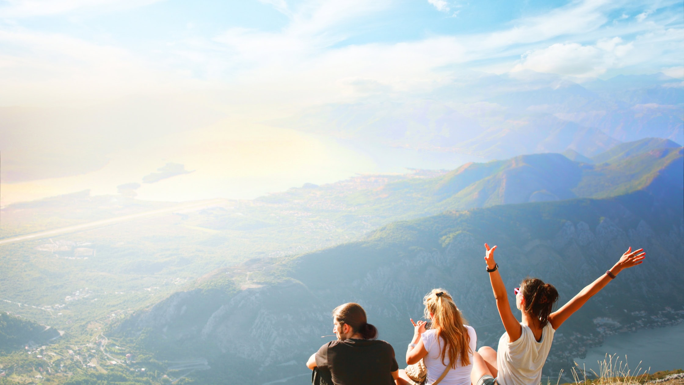 Tres personas se sientan en una montaña con vistas a un pintoresco valle y al agua lejana, y una de ellas levanta los brazos emocionada. El tiempo es claro y soleado. Tres personas se sientan en una montaña con vistas a un pintoresco valle y al agua lejana, y una de ellas levanta los brazos emocionada. El tiempo es claro y soleado.
