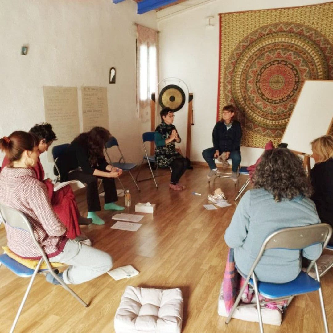 Un grupo de personas sentadas en círculo en sillas y cojines en una sala con suelo de madera, un gong, obras de arte en la pared y una pizarra, participan en un debate o taller.